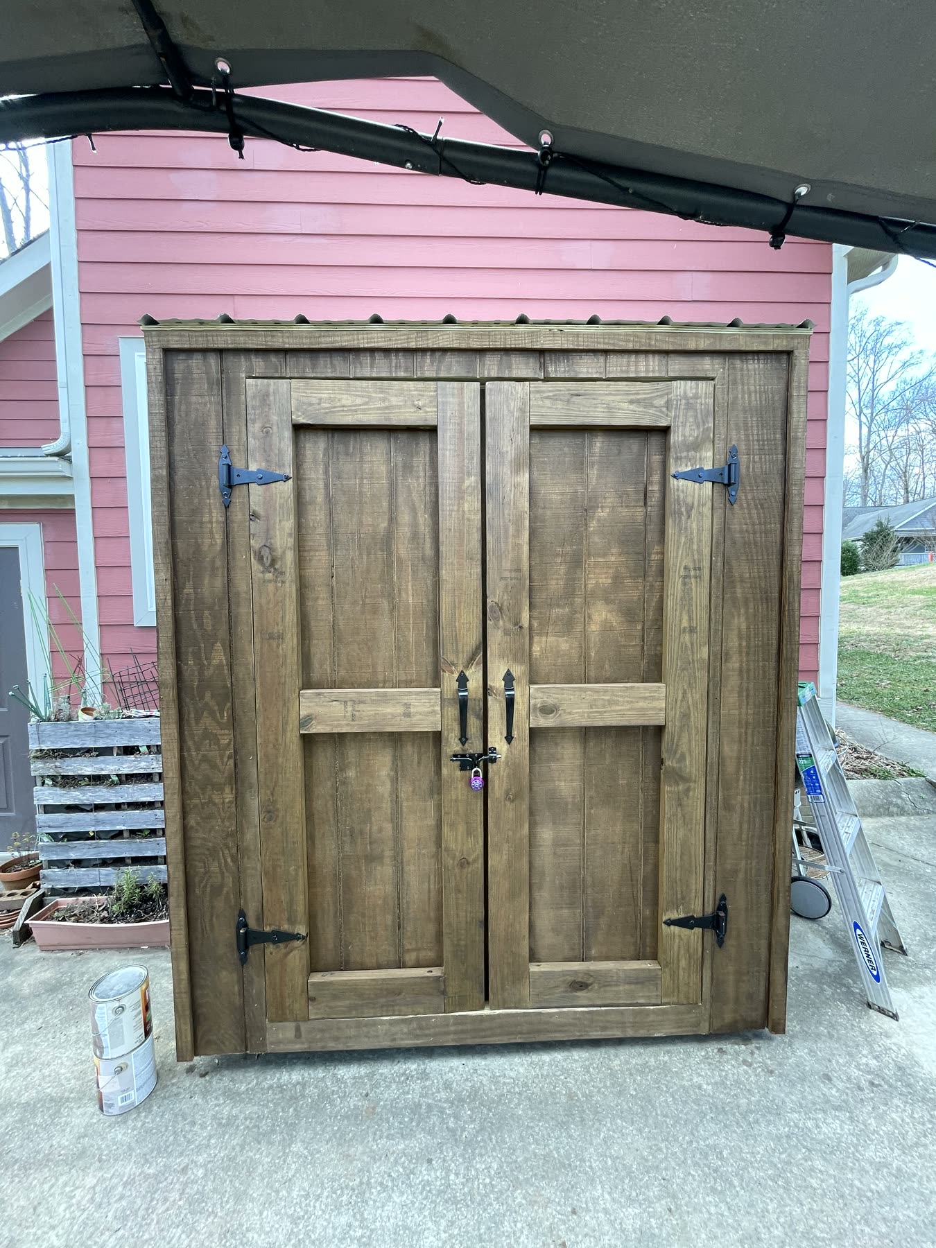 Custom stained-pine storage cabinet with wrought-iron strap hinges against a red clapboard house