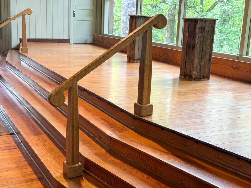 Hand-shaped white-oak scroll handrails on reclaimed plank altar steps in a forest chapel with gable window wall